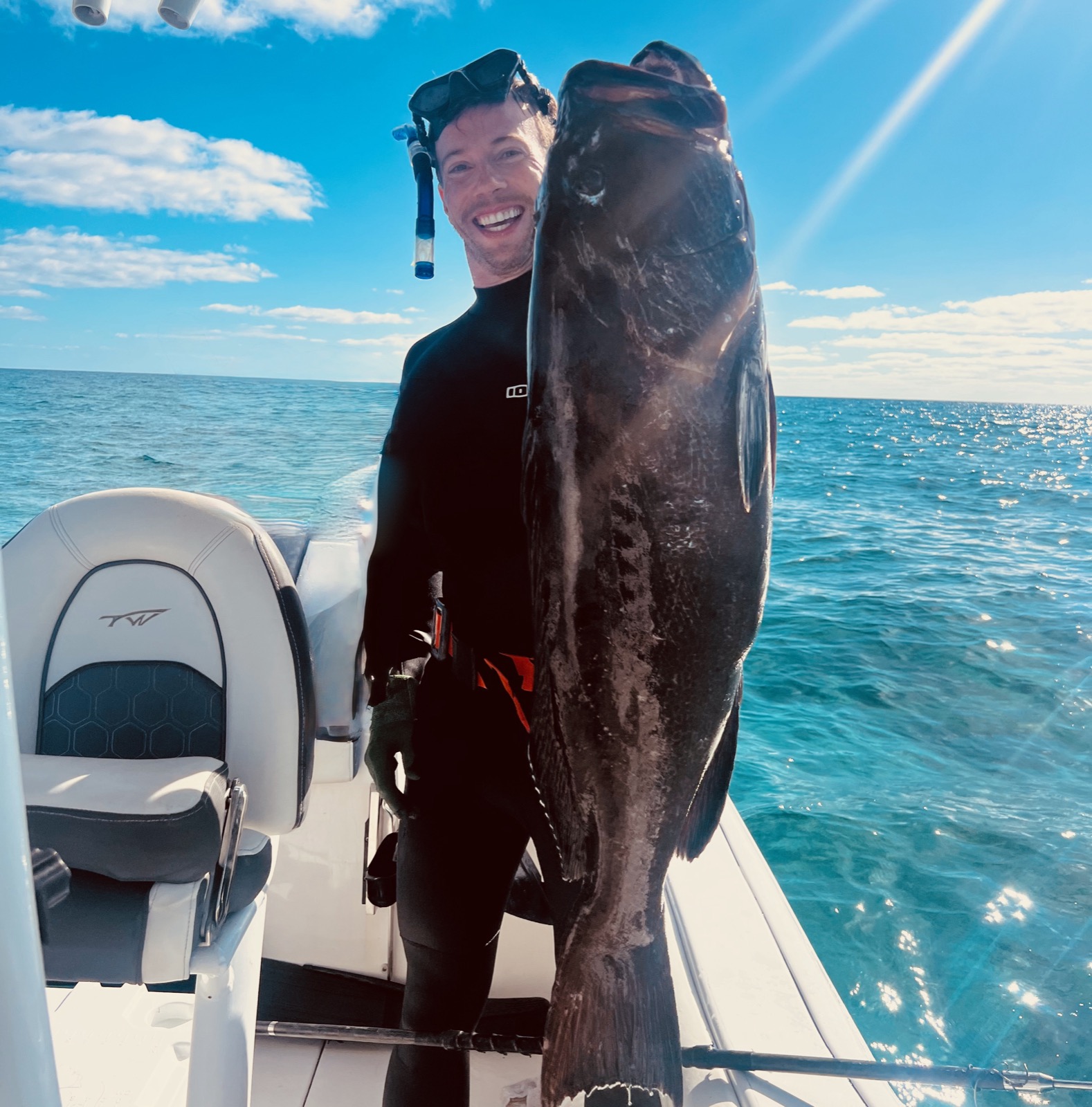 A freediver back on deck holding a big black grouper, mask pushed up, deep blue water behind